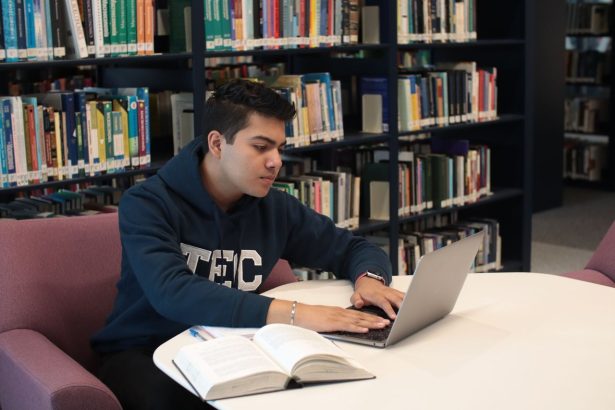 Estudante usa um laptop e consulta livros durante os estudos em uma biblioteca universitária.